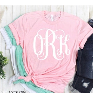 May include: A pink t-shirt with a white monogram "ORK" on the front. The shirt is folded and displayed on a white wooden surface.