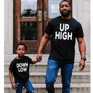 May include: Two people wearing black t-shirts with white text. The adult's shirt says 'Up High' and the child's shirt says 'Down Low'. They are both wearing blue jeans and holding hands while walking down a set of stairs.