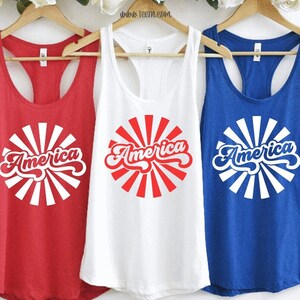 Three racerback tank tops hang together on wooden pole. From left to right is a red, white, and blue racerback tanks. The design is a circle pattern with rays making a circle around the word America. The design is a groovy retro feel.