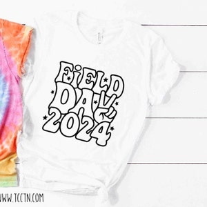 May include: White t-shirt with a black graphic design that says "Field Day 2024" in a retro style font. The graphic includes a peace sign.