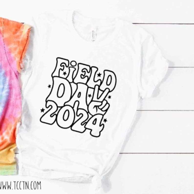 May include: White t-shirt with a black graphic design that says "Field Day 2024" in a retro style font. The graphic includes a peace sign.