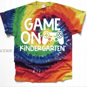 May include: A tie-dye t-shirt with a spiral pattern in rainbow colors. The shirt features the words "GAME ON KINDERGARTEN" in white, along with a white graphic of a video game controller.