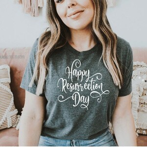 May include: Dark gray t-shirt with a v-neck and the words "Happy Resurrection Day" in white script. The shirt is a short-sleeved tee. The text is in a cursive font.