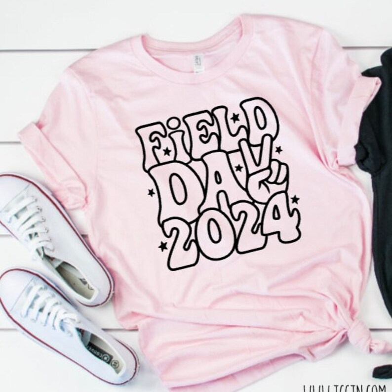 May include: Pink t-shirt with a black graphic that says "Field Day 2024" in a retro font with stars and a peace sign.