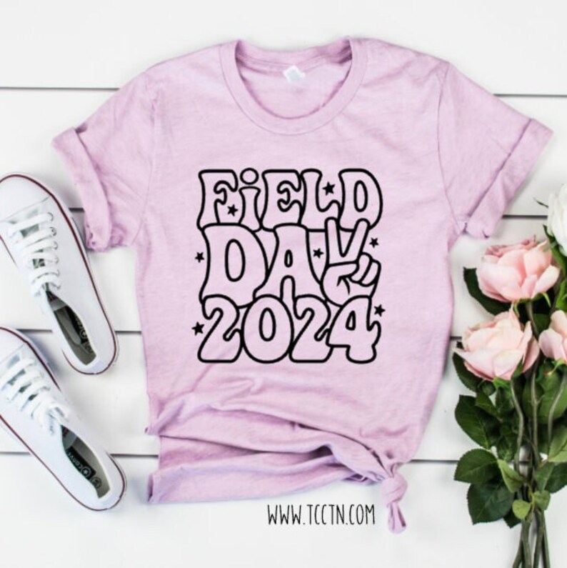 May include: A light purple t-shirt with a black graphic design that says "FIELD DAY 2024" in a retro style font. The graphic includes a peace sign.