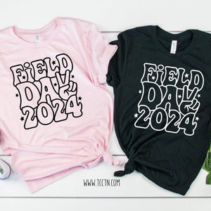 May include: Two t-shirts with a black and white graphic design that says "Field Day 2024" in a retro font. One shirt is pink and the other is black.