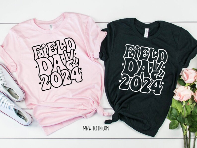 May include: Two t-shirts with a black and white graphic design that says "Field Day 2024" in a retro font. One shirt is pink and the other is black.