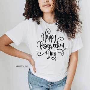 May include: White t-shirt with the words "Happy Resurrection Day" in elegant black script. The shirt is a classic crew neck style and is paired with blue jeans. The website address www.tocti.com is printed on the bottom left of the shirt.