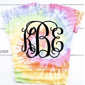 May include: A tie-dye t-shirt with a large black monogram in the centre. The shirt is a rainbow of colours, including pink, orange, yellow, green, and blue.