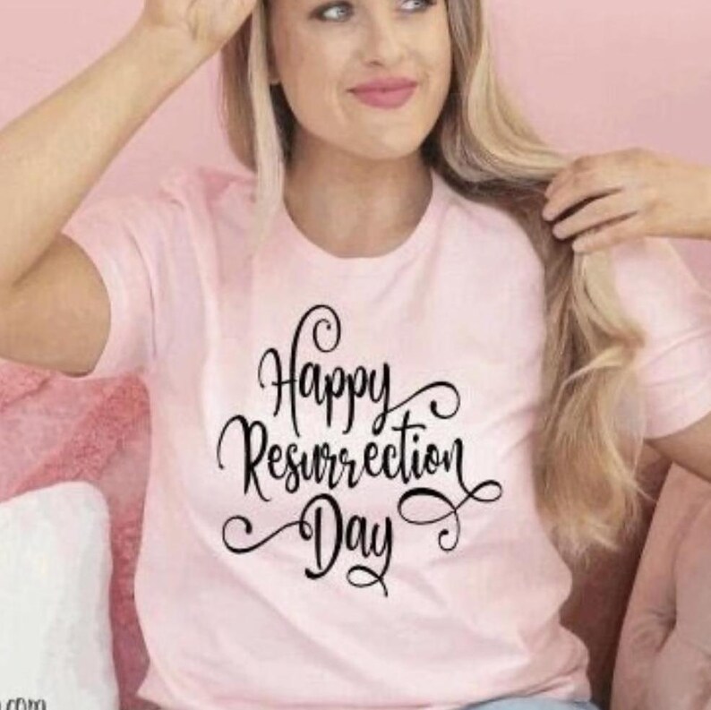 May include: A light pink t-shirt with the words "Happy Resurrection Day" in a black, cursive font. The shirt is a crew neck style and appears to be made of a soft material. The overall theme is Easter.