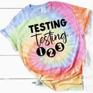 May include: A tie-dye t-shirt with the text "TESTING testing 1 2 3" in black lettering.