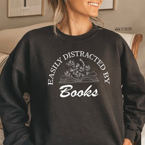 May include: Black sweatshirt with the text "EASILY DISTRACTED BY Books" in white. The design includes an open book with floral accents. The website address www.tcctn.com is printed on the right side of the sweatshirt.