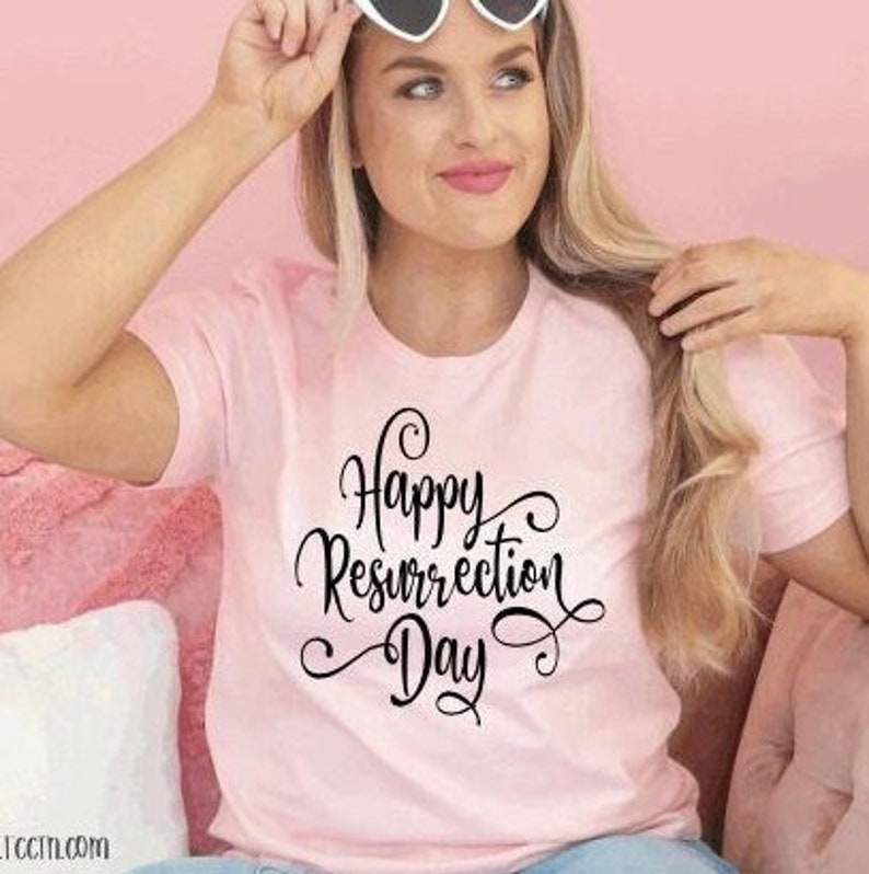 May include: A light pink t-shirt with the words "Happy Resurrection Day" in black script. The shirt is short-sleeved and has a round neckline. The person in the image is wearing heart-shaped sunglasses.