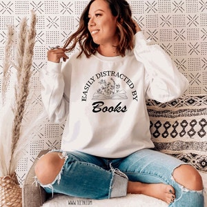 May include: White sweatshirt with the text "EASILY DISTRACTED BY Books" in a black, circular design. The design includes an open book with floral accents. The sweatshirt is long-sleeved and worn with blue jeans.
