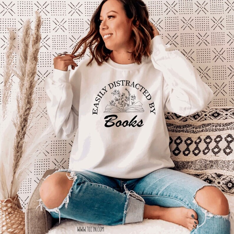 May include: White sweatshirt with the text "EASILY DISTRACTED BY Books" in a black, circular design. The design includes an open book with floral accents. The sweatshirt is long-sleeved and worn with blue jeans.