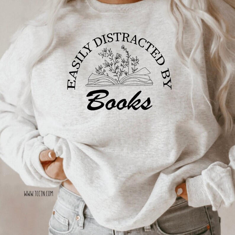 May include: A light gray sweatshirt with the text "EASILY DISTRACTED BY Books" in black. The design includes an open book with floral elements. The sweatshirt is a casual style.