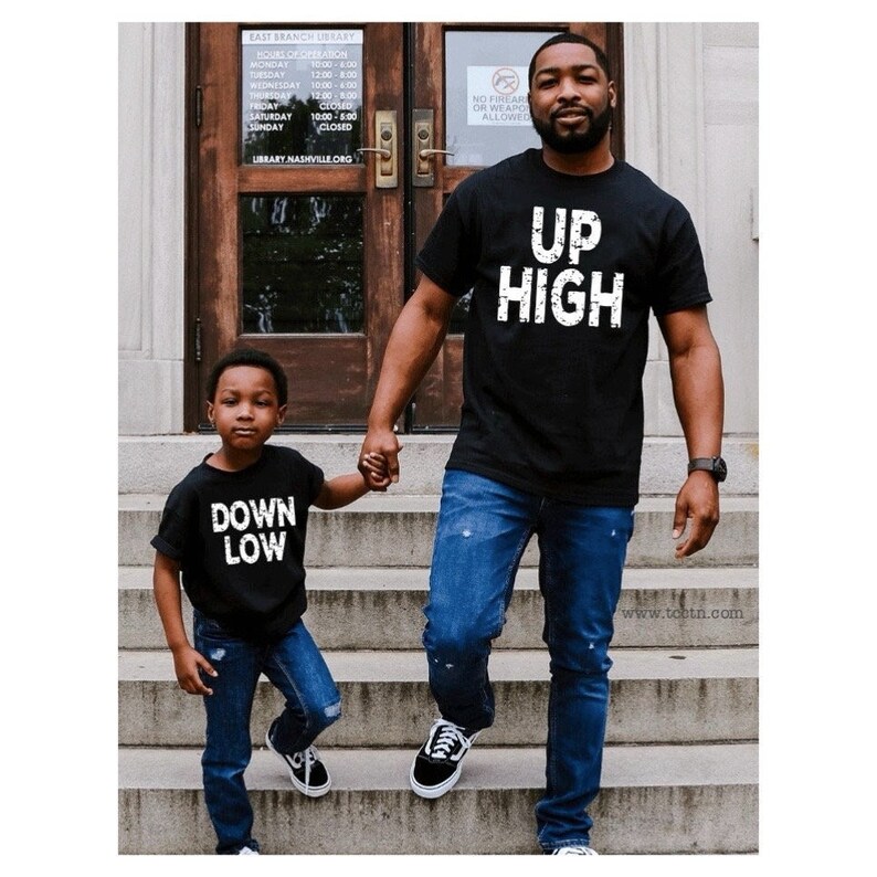 May include: Two people wearing black t-shirts with white text. The adult's shirt says 'Up High' and the child's shirt says 'Down Low'. They are both wearing blue jeans and holding hands while walking down a set of stairs.