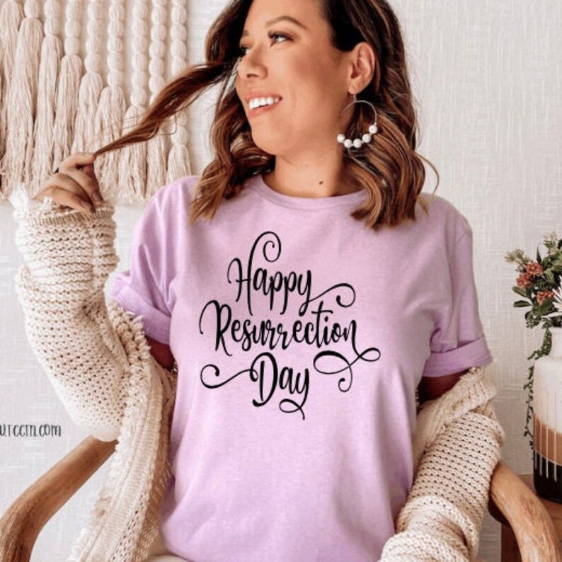 May include: A light purple t-shirt with the words "Happy Resurrection Day" in black script. The shirt has rolled-up sleeves and is being worn by a person. The person is wearing large hoop earrings with white pearls.