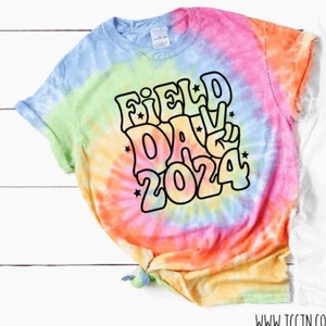 May include: A tie-dye t-shirt with a rainbow swirl pattern. The shirt has a black outline of the text "FIELD DAY 2024" in a retro font.