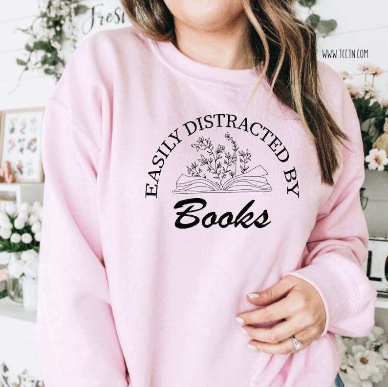 May include: A light pink sweatshirt with the text "EASILY DISTRACTED BY Books" in black. The graphic features an open book with floral elements. The sweatshirt is a casual, comfortable style.