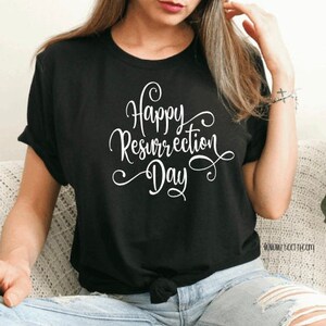 May include: Black t-shirt with the white text "Happy Resurrection Day" in a cursive font. The shirt is a crew neck and has short sleeves. The shirt is tied at the waist.