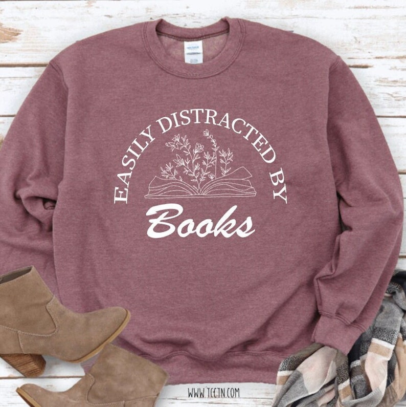 May include: A maroon sweatshirt with the text "EASILY DISTRACTED BY Books" in a white, circular design. The design includes an open book with floral elements. The bottom of the sweatshirt shows a pair of brown boots and a scarf.