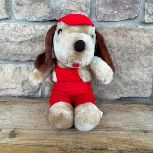 Puede incluir: Un peluche de perro con pelaje beige, orejas marrones y nariz negra. El perro lleva una gorra roja y un peto rojo. El juguete está sobre una superficie de madera. La boca del perro es roja.