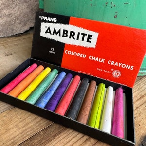 Può includere: Una scatola aperta di pastelli a cera colorati Prang Ambrite. La scatola ha un design rosso e nero con la scritta "Colored Chalk Crayons". All'interno ci sono 12 pastelli colorati in vari colori, tra cui rosa, arancione, giallo e blu.