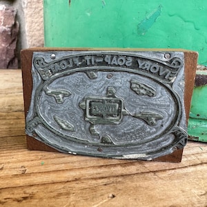 Puede incluir: Un sello de metal vintage con el texto "SAVORY SOAP-IT-FLOATS" en un marco ovalado. El sello está montado sobre un bloque de madera. El metal tiene un diseño detallado y ornamentado.