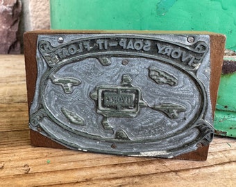Puede incluir: Un sello de metal vintage con el texto "SAVORY SOAP-IT-FLOATS" en un marco ovalado. El sello está montado sobre un bloque de madera. El metal tiene un diseño detallado y ornamentado.