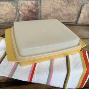 May include: A yellow square Tupperware container with a matching lid. The lid is a light beige color and sits on top of the yellow base. The container is resting on a striped towel with brown, pink, and green stripes.