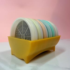 May include: A set of colorful coasters in a yellow holder. The coasters are pastel shades of white, yellow, peach, and blue. The holder has a curved design and is made of yellow plastic.