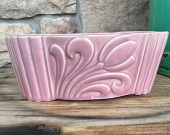 May include: A rectangular, light pink ceramic planter with a tulip design. The planter has a ribbed texture on the sides and a curved shape. The tulip design is raised and detailed.