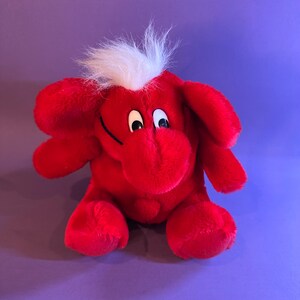 May include: A red plush toy with white fur on its head. The toy has large, white eyes and a black nose. It is sitting on a purple background.