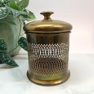 May include: A vintage brass container with a lid. The container has a decorative openwork design around the middle. The lid has a small knob on top. The container is cylindrical and sits on a white surface.