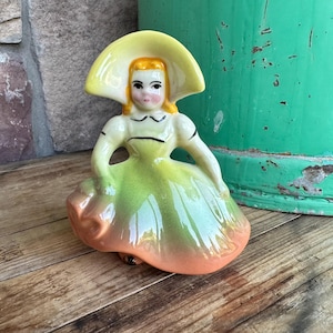 Könnte beinhalten: Vintage Keramikfigur einer Frau mit breitkrempigem Hut und langem Kleid. Das Kleid wechselt von Grün zu Orange, und der Hut ist gelb. Die Figur hat bemalte Gesichtszüge und sitzt.