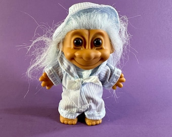 May include: A vintage Troll doll with a smiling face, wearing a blue and white striped pajama set and nightcap. The doll has light blue hair and a white bow at the waist. The doll is standing on a purple surface.