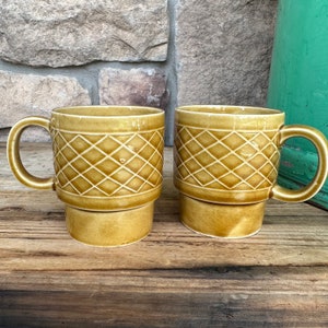 May include: Two matching ceramic mugs in a warm, golden-brown colour, featuring a diamond pattern. Each mug has a rounded handle and a pedestal base. The mugs are displayed on a wooden surface.