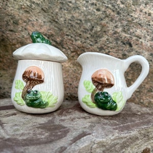 May include: A cream-colored ceramic sugar bowl with a mushroom-shaped lid and a matching creamer. Both pieces are decorated with a brown and tan mushroom, a green frog, and green leaves. The sugar bowl lid has a green frog on top.