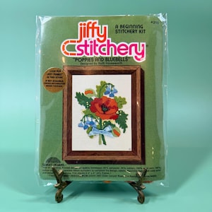May include: A Jiffy Stitchery Poppies and Bluebells beginning stitchery kit in a clear plastic package. The packaging features a green background with a printed image of a floral embroidery design in a brown frame and the text Jiffy Stitchery.