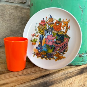 Puede incluir: Un plato blanco con una colorida ilustración de un tren que transporta animales de dibujos animados, como un oso, un león y una jirafa. A la izquierda del plato hay un vaso de plástico naranja. El plato tiene las palabras "Toyland Express" y "Toyland".