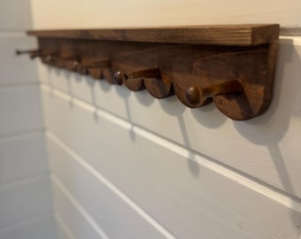 May include: A long, dark brown wooden wall-mounted rack featuring a flat top shelf and a series of rounded, scalloped edges along the bottom. Multiple wooden pegs extend outward from the scalloped base, providing hooks for hanging various items.