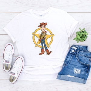 May include: White t-shirt with a cartoon image of Woody from Toy Story. Woody is wearing his signature cowboy hat and vest, and he is standing in front of a sheriff's star.