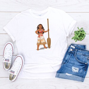 May include: White t-shirt featuring a cartoon illustration of a Polynesian woman holding a wooden paddle. The woman is wearing a brown top, a grass skirt, and a necklace.