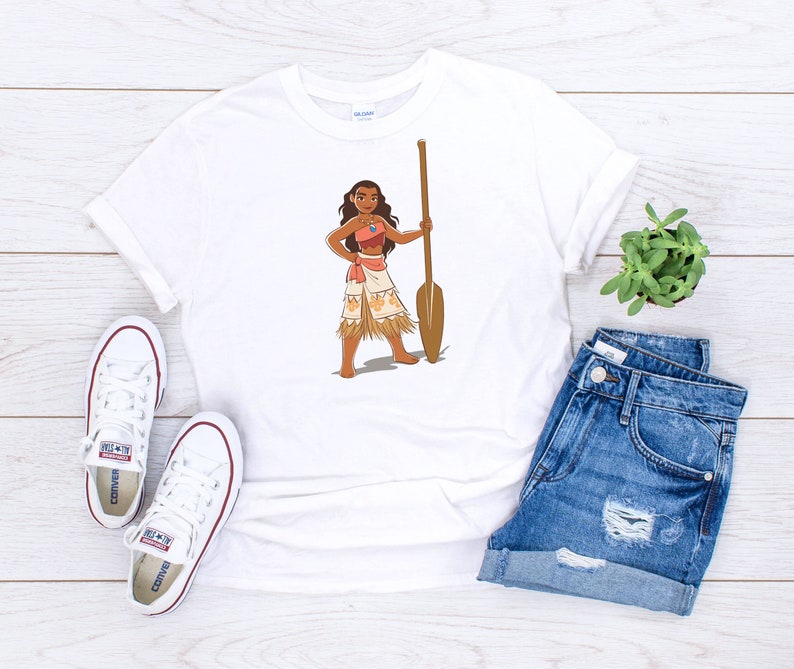 May include: White t-shirt featuring a cartoon illustration of a Polynesian woman holding a wooden paddle. The woman is wearing a brown top, a grass skirt, and a necklace.
