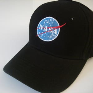 May include: Black baseball cap featuring the NASA logo. The logo is a blue circle with white stars, the word "NASA" in white, and a red swoosh. The cap has a curved brim and a structured fit.
