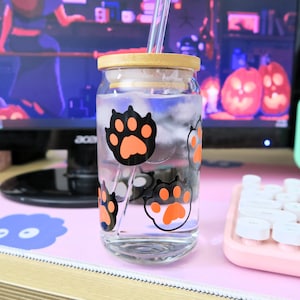 May include: A clear glass jar with a wooden lid and a straw. The jar has orange paw prints on it and is filled with water. The jar is sitting on a pink and white desk mat.