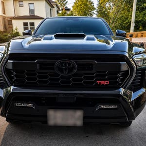 Op de afbeelding: Een zwarte Toyota Tacoma pick-up truck gezien vanaf de voorzijde, met een opvallende zwarte honingraatgrille met een centraal Toyota-embleem en een rood TRD-badge. Het voertuig heeft een motorkap met luchthapper, hoekige koplampen en geïntegreerde bumperverlichting.