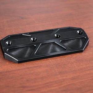 May include: A black metal component with a geometric design. The component has a rectangular shape with beveled edges and several circular holes. The surface has a textured, brushed appearance. The item is displayed on a wooden surface.