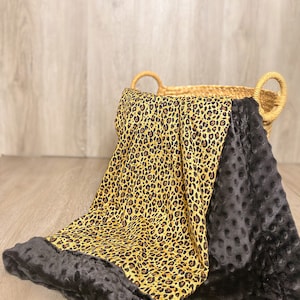 May include: A soft, plush blanket with a leopard print on one side and black minky fabric on the other side. The blanket is folded and placed in a woven basket.